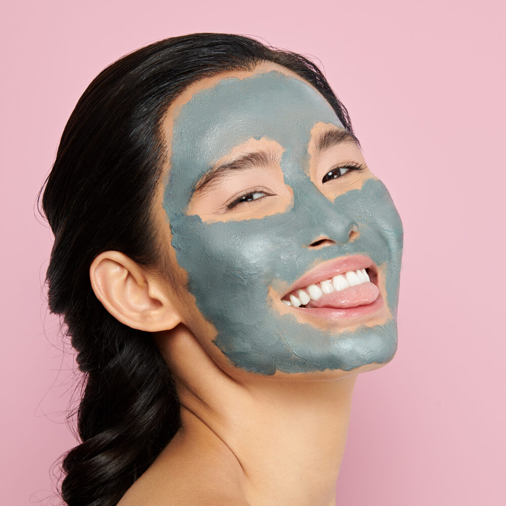 A woman with black hair and a green mask treatment on her face smiles at the camera while sticking her tongue out through her teeth.