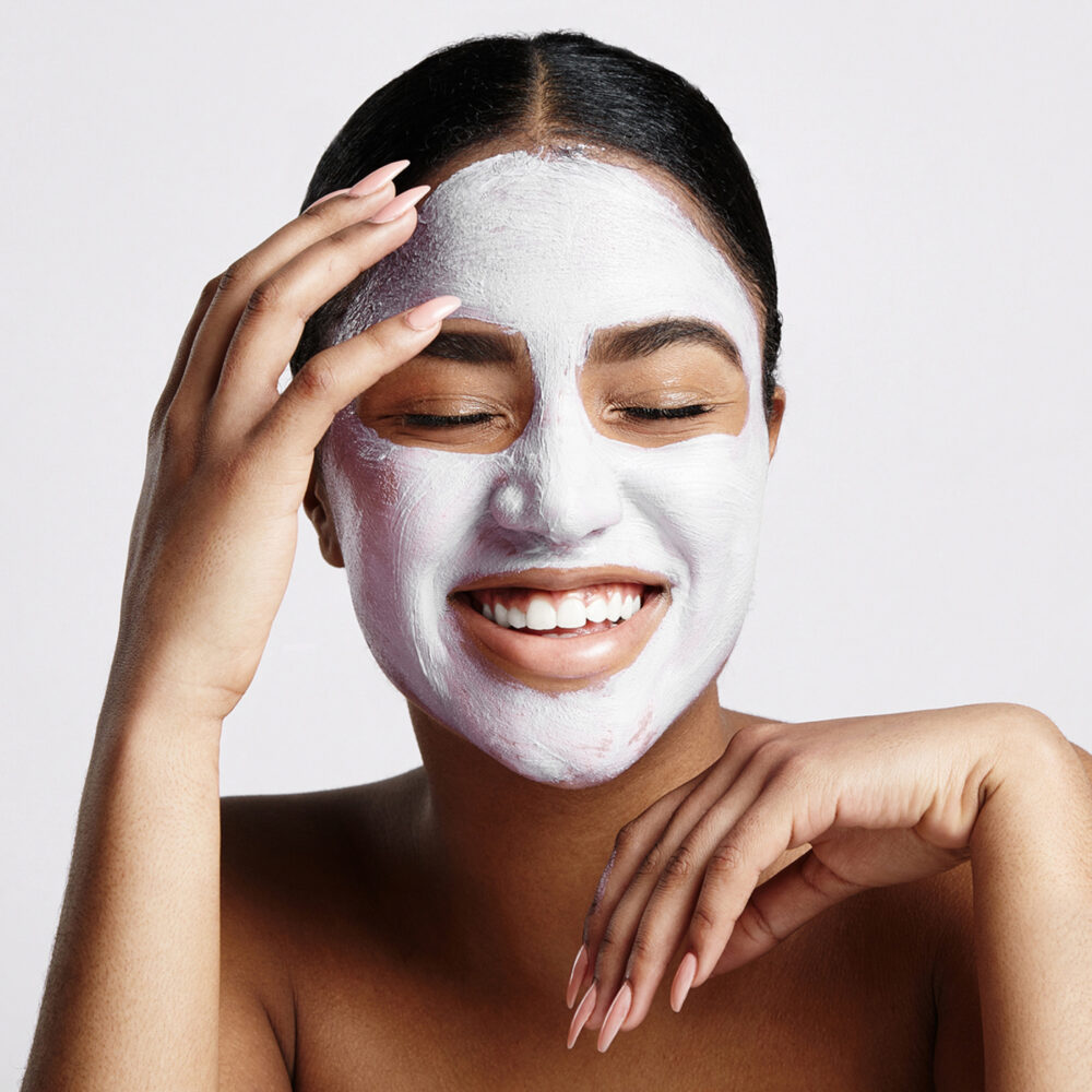 A white woman with dark hair smiles with her eyes closed and her hands around her face. A white facial mask has been applied to her face.