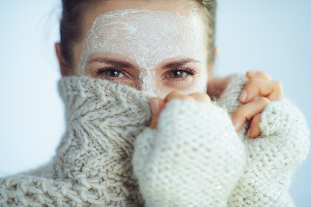 elegant woman in roll neck sweater and cardigan with white facial mask hiding behind clothes on winter light blue background.