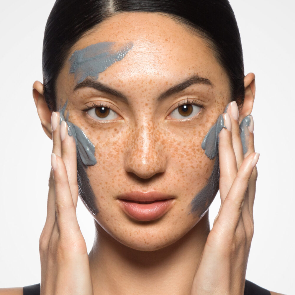 A woman with black hair looks directly at the camera while applying a green mask to her face with her hands.