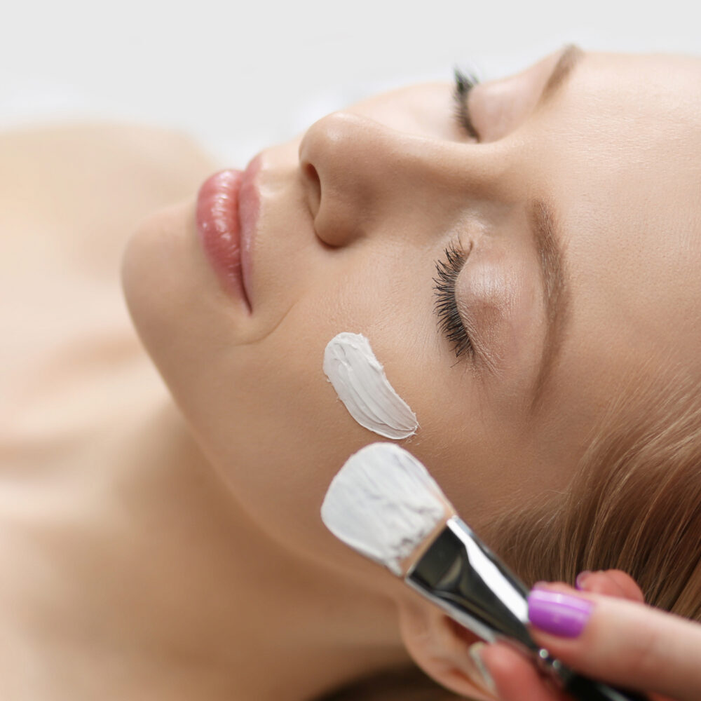 A white woman with light hair closes her eyes and smiles as a facial treatment is applied with a brush.