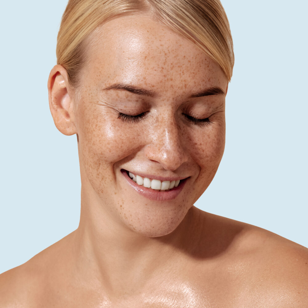 A white woman with light hair and freckles smiles with both eyes closed.